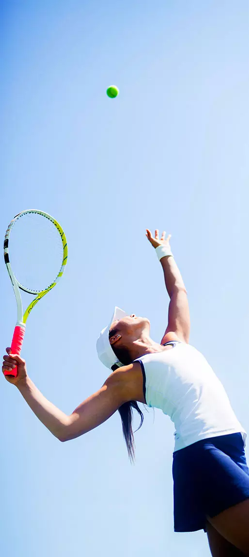 Giocatrice di tennis che serve la palla sotto un cielo azzurro, racchetta in primo piano e cappellino bianco.