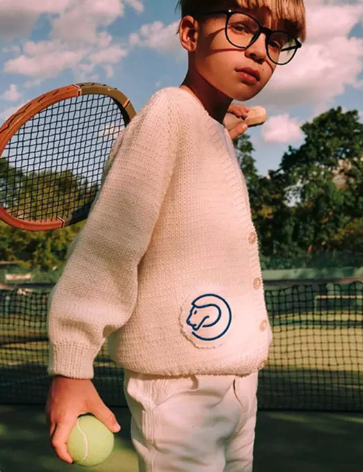 bambino in una maglia beige con patch circolare blu sul lato, in campo da tennis accanto alla rete