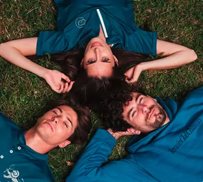 Tre amici sdraiati sull'erba, testa in centro, cercano di guardare la fotocamera con espressioni rilassate e sorrisi.
